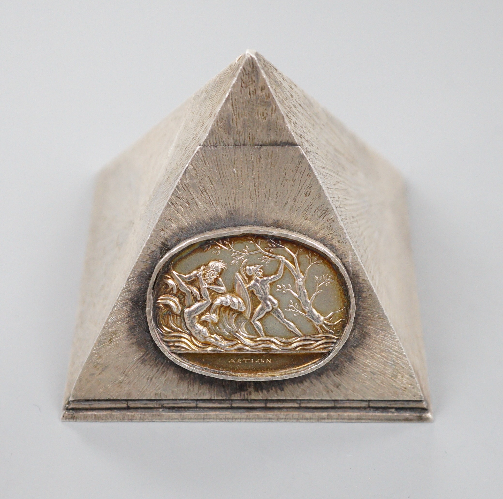 A textured white metal pyramid shaped box, the hinged panel decorated with Greek scene and containing five dice, height 62mm.